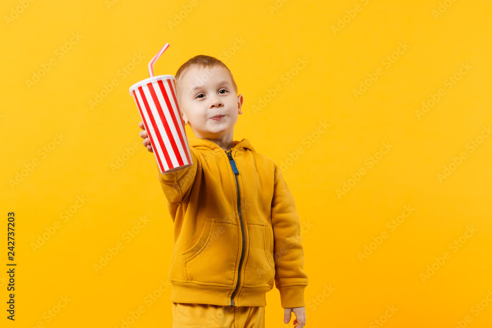 Fototapeta premium Little fun cheerful kid boy 3-4 years old wearing yellow clothes hold cup of soda isolated on orange wall background, children studio portrait. People, childhood lifestyle concept. Mock up copy space.