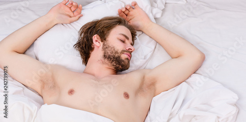 Picture showing young man stretching in bed. Young man stretching while waking up in the morning. Man with eyes still closed reaching button on the clock to turn it off.