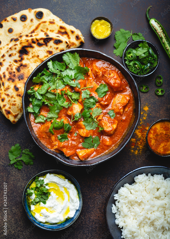 Traditional Indian dish Chicken tikka masala with spicy curry meat in bowl, basmati rice, bread naan, yoghurt raita sauce on rustic dark background, top view, close up. Indian style dinner from above