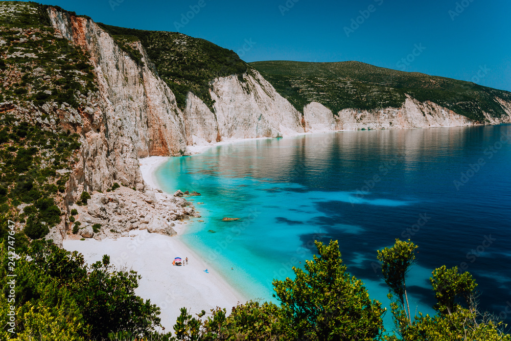 Fototapeta premium Amazing Fteri beach lagoon, Kefalonia, Greece. Tourists under umbrella chill relax near clear blue emerald turquoise sea water