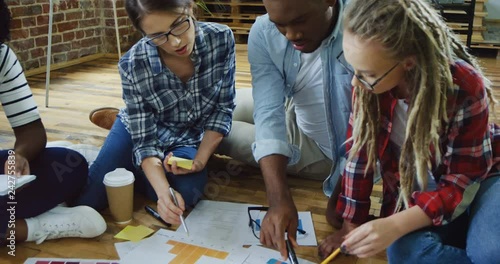 Close up of the mixed-races team of young men and women sitting on the floor and working over the common sturtup business plan with graphics and charts. Indoors. photo