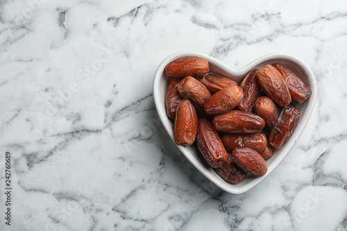 Heart shaped bowl with sweet dried date fruits on marble background, top view. Space for text