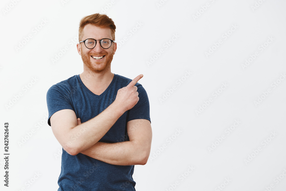 Cute and friendly businessman showing his office and projects to friend. Portrait of handsome redhead male with bristle in glasses and t-shirt, pointing at upper right corner and smiling joyfully Cute and friendly businessman showing his office and projects to friend. Portrait of handsome redhead male with bristle in glasses and t-shirt, pointing at upper right corner and smiling joyfully