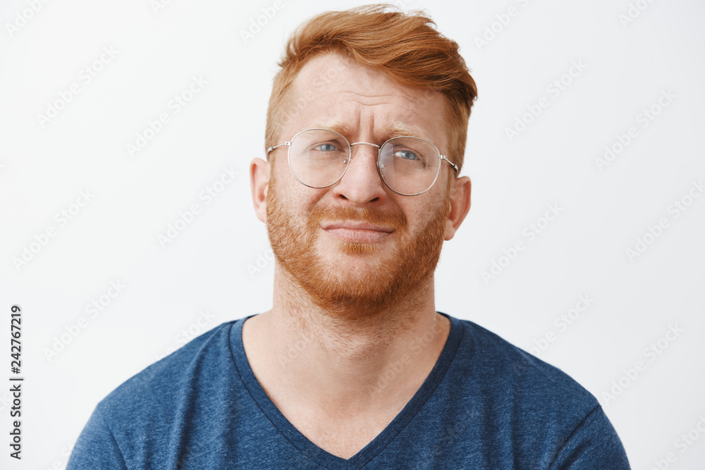 Obraz premium Handsome gloomy redhead male with beard frowning and pursing lips looking right with regret and disappointment, standing over gray background in casual clothes and glasses