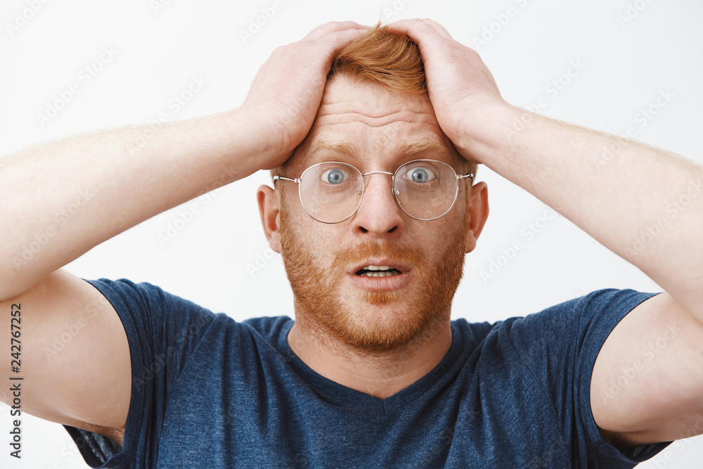 Headshot of worried attractive redhead businessman in glasses and blue t-shirt holding hands on hair and frowning, standing in stupor at shocking scene, feeling anxious and desperate Headshot of worried attractive redhead businessman in glasses and blue t-shirt holding hands on hair and frowning, standing in stupor at shocking scene, feeling anxious and desperate