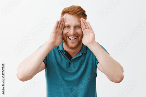 Studio shot of playful and joyful good-looking young redhead father with bristle holding palms along face and smiling broadly, playing peekaboo with kids and having fun in family circle over gray wall photo