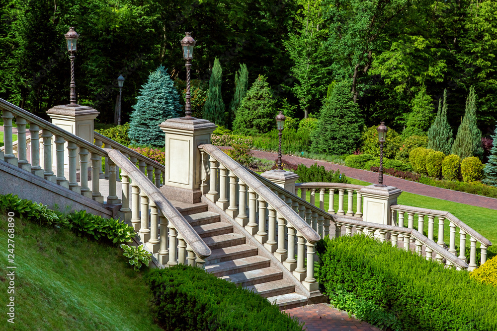 a stone staircase with railings and balustrades on pedestals are installed iron lantern pillars with lanterns in a park with greenery and landscape design in the form of hedges of bushes and trees. a stone staircase with railings and balustrades on pedestals are installed iron lantern pillars with lanterns in a park with greenery and landscape design in the form of hedges of bushes and trees.