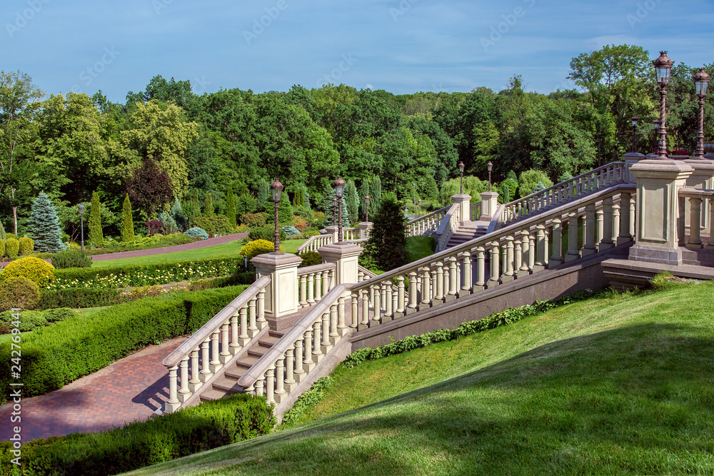 A staircase with stone railing balustrades and retro lanterns on pedestals against the backdrop of a landscape with a boxwood hedge and a forest with trees.