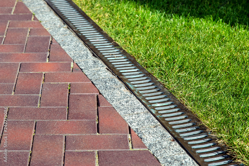 Drainage system with iron mesh along the pedestrian sidewalk with red tiles and green lawn, close-up of iron rainfall. Drainage system with iron mesh along the pedestrian sidewalk with red tiles and green lawn, close-up of iron rainfall.