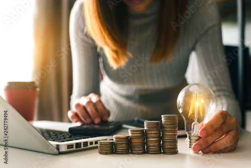business hand holding lightbulb with using laptop computer and money stack in office. idea saving energy and accounting finance concept in morning light