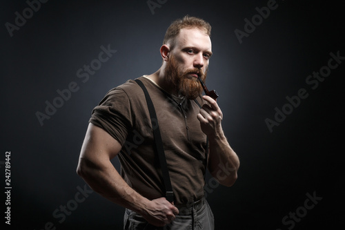 Lumberjack brutal red beard muscled man in brown shirt with smoking tube standing on dark background
