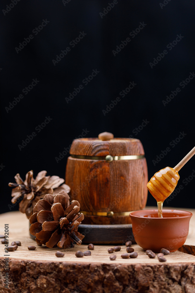 A barrel with honey and a clay cup with a honey stick on a large wooden saw cut surrounded by pine cones on a dark background. Copy space. Space under your text.