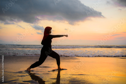 silhouette of young fit Muslim woman covered in Islam hijab head scarf training martial arts karate punch attack and fitness workout at beautiful beach sunset