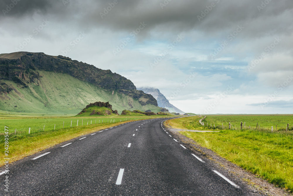 Naklejka premium Asphalt road in Iceland countryside among green fields and rocky mountains on cloudy summer day. Travel on Ring Road number 1