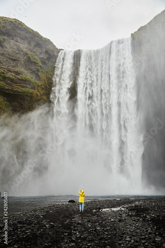A little girl in a yellow raincoat stands in front of a huge waterfall. Iceland.