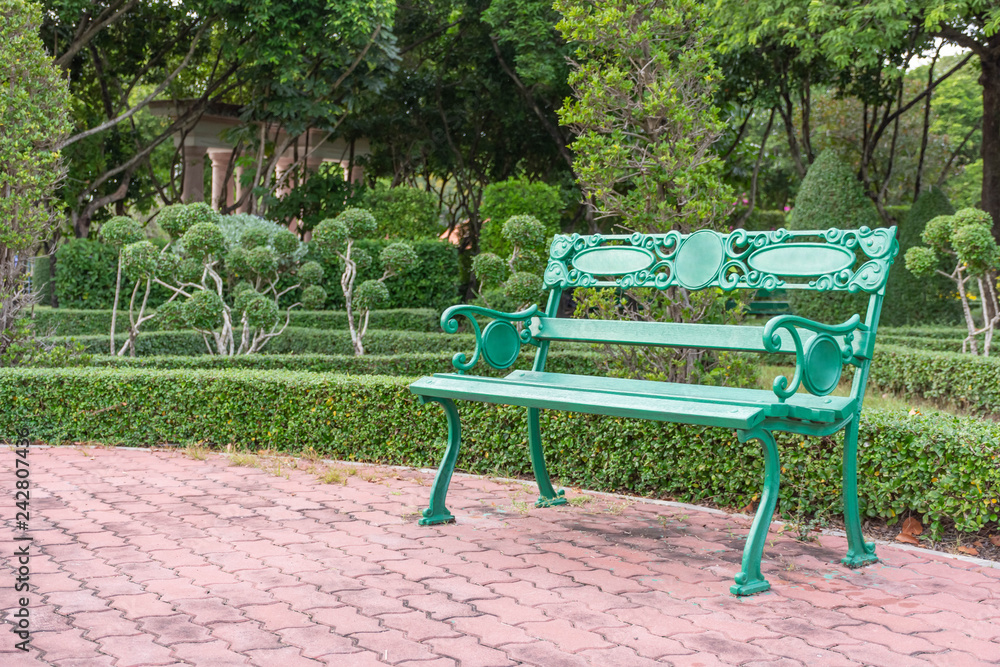 One of the green benches located on the patio, brick brown blog. In a park with lots of trees,