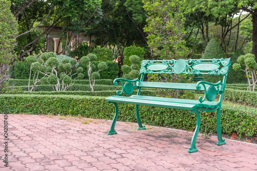 One of the green benches located on the patio, brick brown blog. In a park with lots of trees,