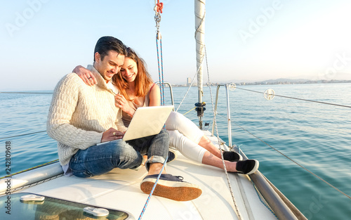 Young couple in love on sail boat having fun remote working at laptop- Happy luxury lifestyle on yacht sailboat - Technology concept with influencer travel blogger - Warm afternoon color tone filter