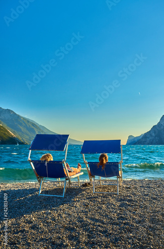 People sitting at the beach on the lounge chair and admiring Lake Garda in the summer time View of the beautiful Lake Garda surrounded by mountains