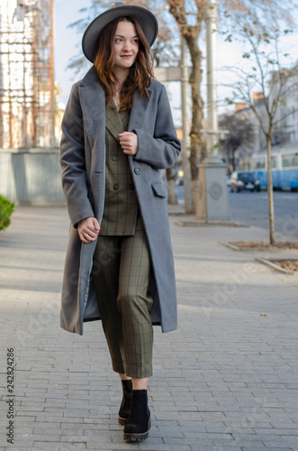 Outdoor portrait of young beautiful fashionable happy lady posing on a street of the old city. Model wearing stylish wide-brimmed hat a women's suit with trousers and autumn coat. 