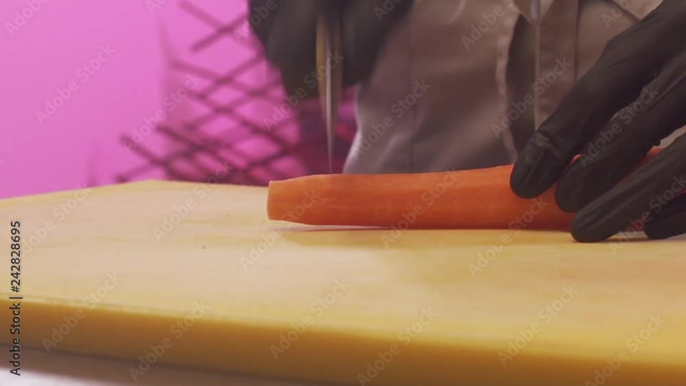 Closeup The Hand Of A Cook In Protective Gloves Cuts A Carrot By A Large Knife On A Wooden Cutting Board. Concept Of Restaurant Cuisine. Slow Motion Closeup The Hand Of A Cook In Protective Gloves Cuts A Carrot By A Large Knife On A Wooden Cutting Board. Concept Of Restaurant Cuisine. Slow Motion