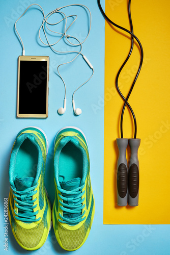 Pair of sport shoes, skipping rope and cellphone with earphones. New sneakers on blue and yellow pastel background, copy space. Overhead shot of running foot wear. Top view, flat lay