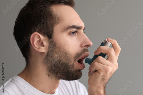 Young man with inhaler having asthma attack on grey background