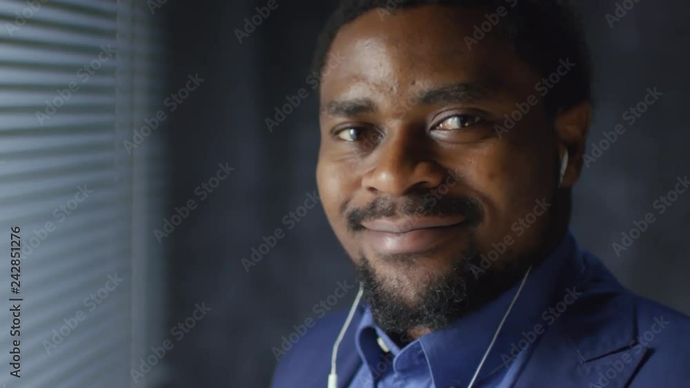Portrait of bearded black man standing by window with slatted venetian blinds on it, listening to music with earphones, then looking at camera Portrait of bearded black man standing by window with slatted venetian blinds on it, listening to music with earphones, then looking at camera