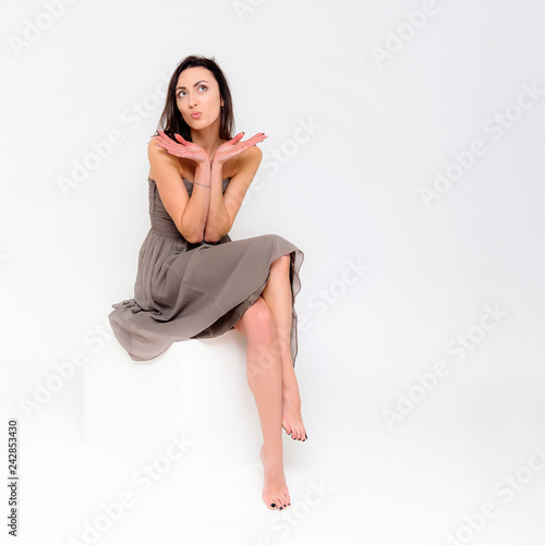 Concept portrait of a brunette girl, sitting on a white background in a beautiful brown dress on a white cube in studio. She sits right in front of the camera in various poses with different emotions.
