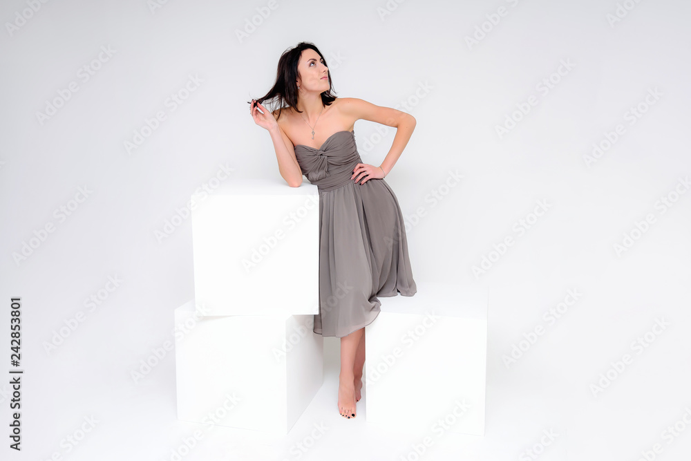 Concept portrait of a brunette girl, standing on a white background in a beautiful brown dress near the white cube. She sits right in front of the camera in various poses with different emotions. Concept portrait of a brunette girl, standing on a white background in a beautiful brown dress near the white cube. She sits right in front of the camera in various poses with different emotions.