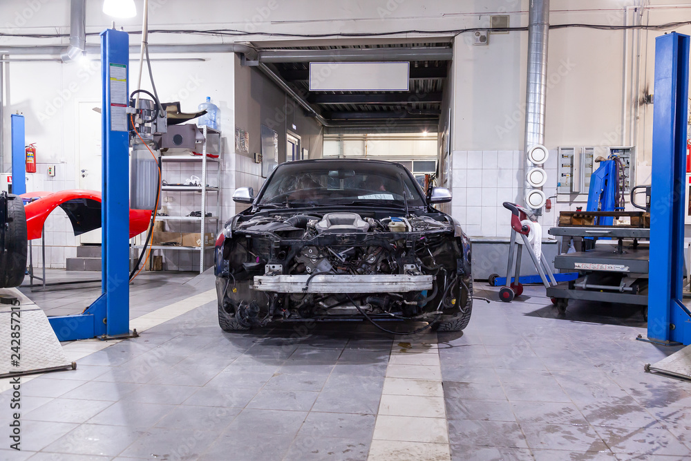 Disassembled black car in the workshop near the lift cars prepared for repair: front end, hood and bumper removed, internal equipment, parts and spare parts visible.