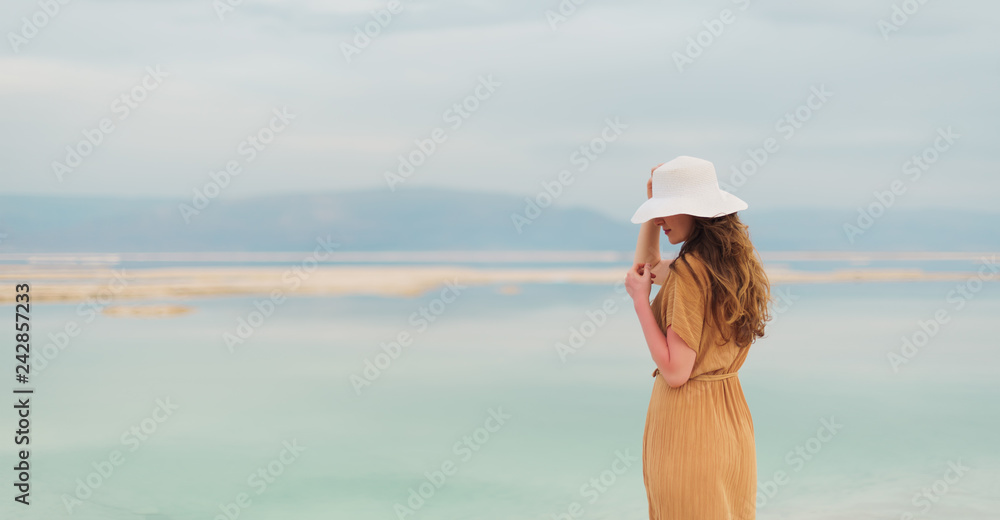 Back view of stylish girl wearing dress on seaside, Dead Sea beach. Travel, summer vacation, holiday, freedom concept. Digital detox. Sea background with copy space. Banner.