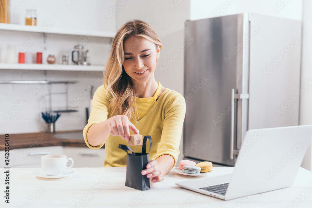 attractive woman putting small paper box into black shopping bag in kitchen attractive woman putting small paper box into black shopping bag in kitchen