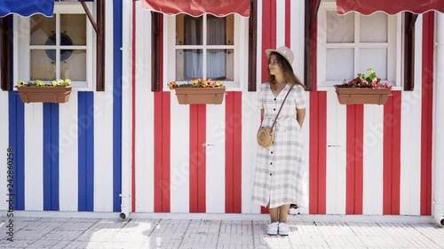 Pretty woman in checkered dress and hat smiling and enjoying sunny day. Girl standing at background striped facades on street of Costa Nova, Portugal