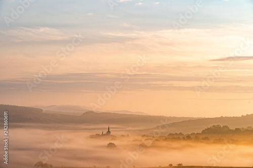 View of village covered in fog during morning sunrise. South moravian landscape with low clouds during a sunrise. Hazy summer scene of small village.