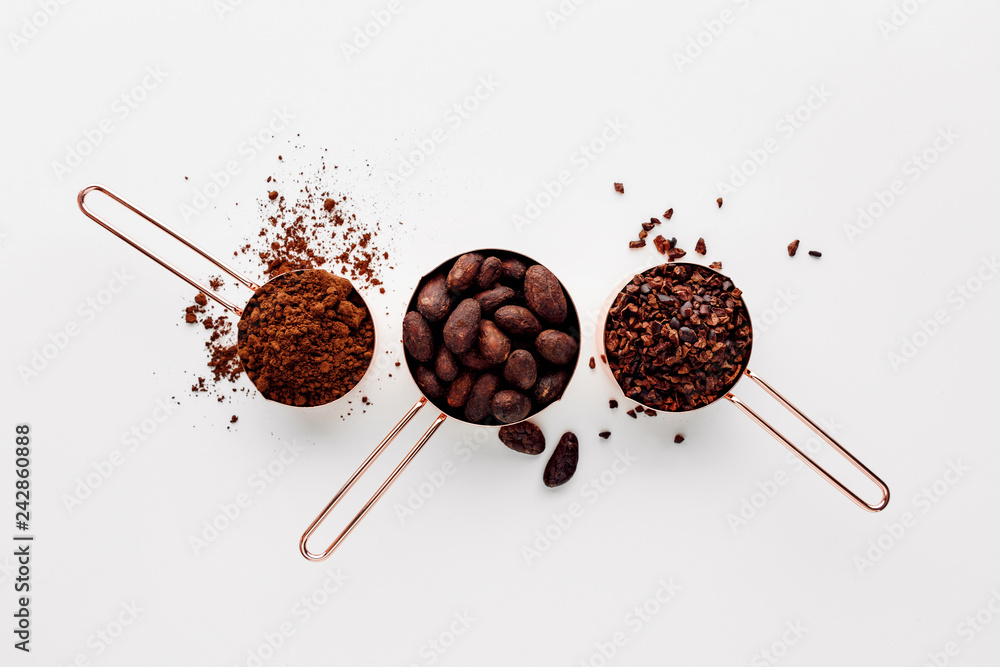 Rose gold measuring cups of cocoa beans, cacao nips and cocoa powder on a white background, flat lay healthy food concept - obrazy, fototapety, plakaty 