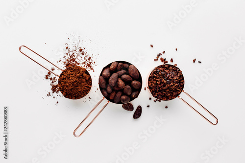 Rose gold measuring cups of cocoa beans, cacao nips and cocoa powder on a white background, flat lay healthy food concept