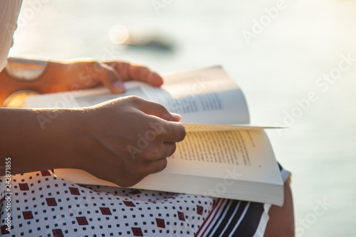 Young woman reading book at sunset in front of the sea on Ponza island coast, sitting on a wall with view of the ocean.