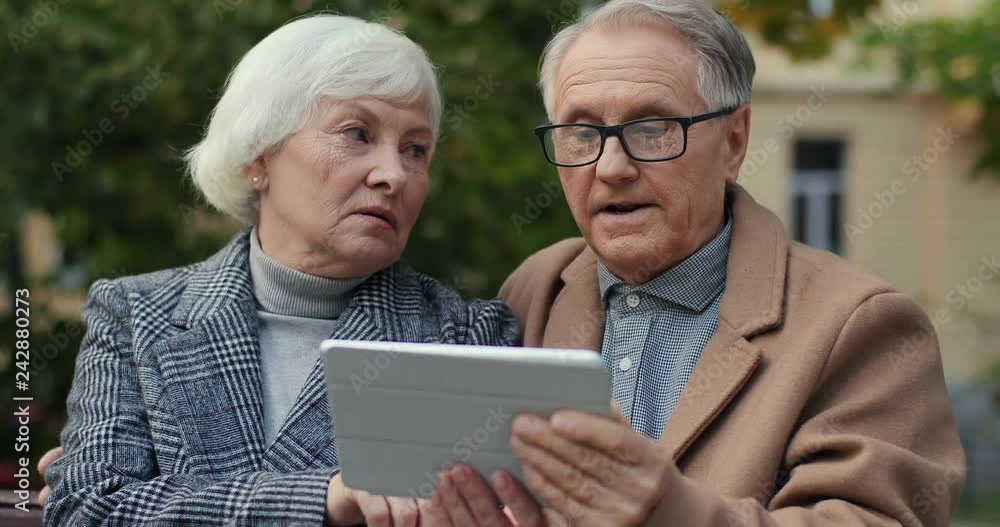 Portrait of the old Caucasian man and woman sittingon the bench and watching something on the tablet device. At fall. Outside. Portrait of the old Caucasian man and woman sittingon the bench and watching something on the tablet device. At fall. Outside.