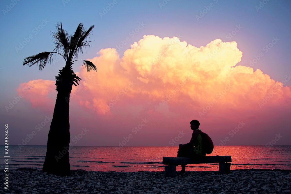 Silhouette girl with backpack sitting on bench on an empty beach with lonely palm tree and looking at an amazing purple sunset. Inspirational moment