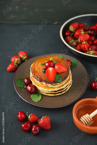 pancakes in a plate with strawberries, mint, honey and cherry on a dark black background.