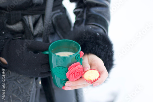 A girl in winter clothes is drinking coffee from a mug. In his other hand holds a cupcake. Outdoor picnic