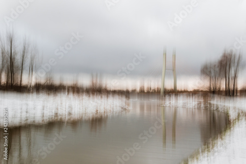 Blurred winter landscape with trees, bridge pilier, snow and reflections in water at sunset
