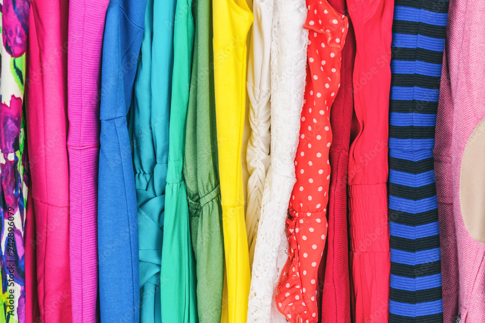 Closet women's fashion outfits clothes arranged in rainbow colors assorted. Clothing store dresses hanging on shopping rack. Variety of fabrics and patterns, wool, polyester, polka dots, stripes.