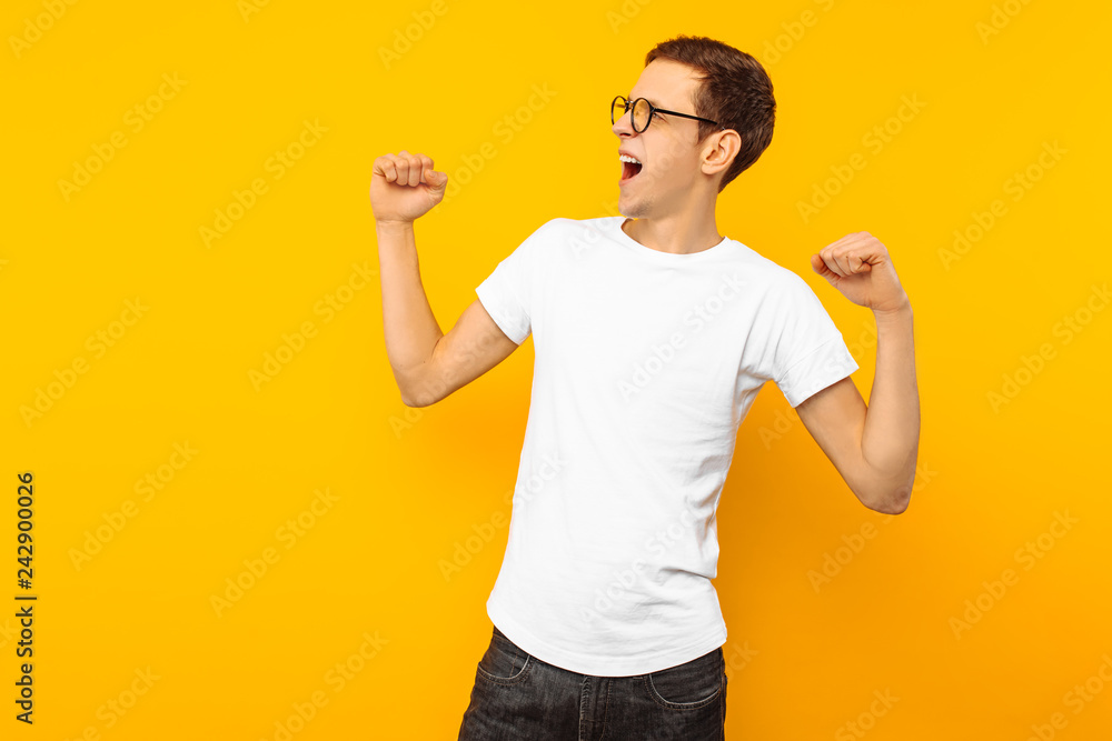 man in glasses, dressed in a white T-shirt, the man who won shows a gesture of victory and success, on a yellow background man in glasses, dressed in a white T-shirt, the man who won shows a gesture of victory and success, on a yellow background