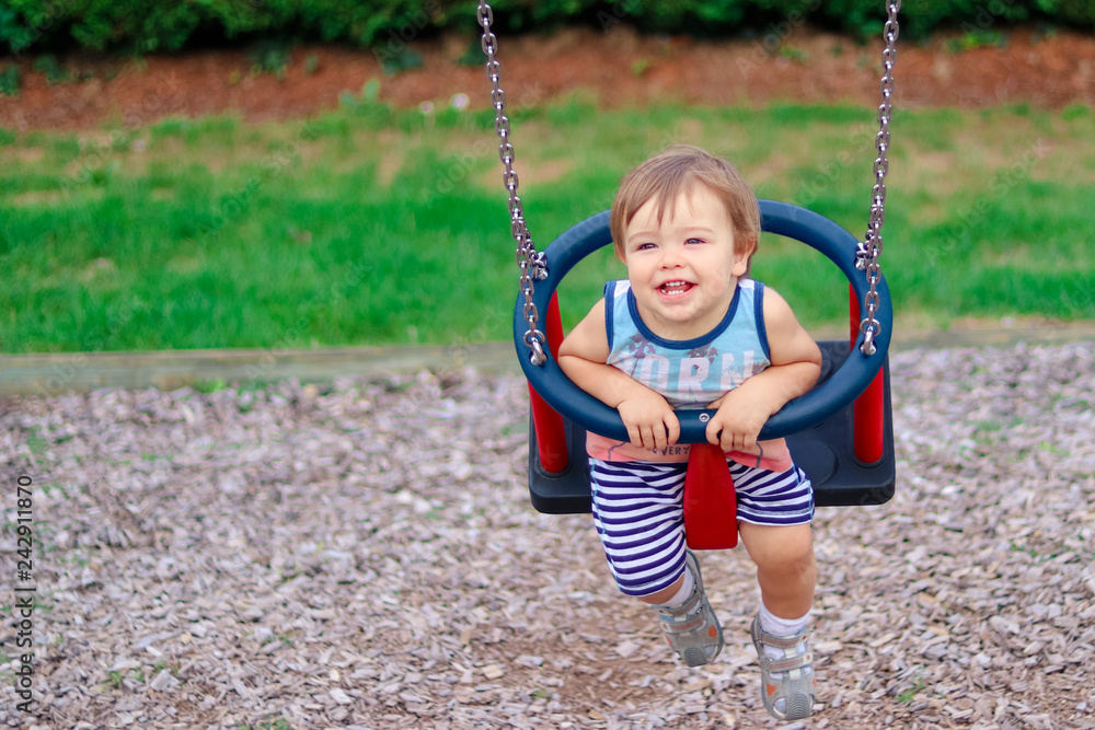 Cute happy laughing little boy having fun riding on swing at children playground on summer day with open mouth showing his teeth. Carefree childhood. Copy space.