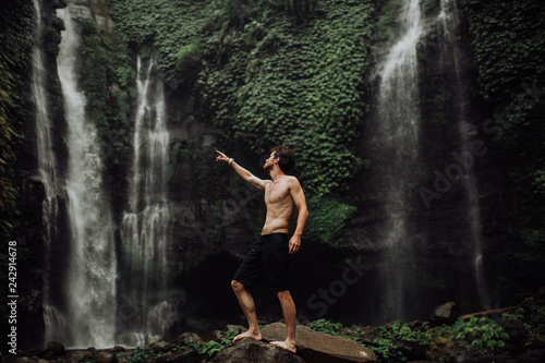 young man muscular and beautiful standing near the waterfall and shows forward