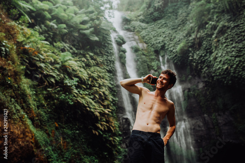 Fototapeta Naklejka Na Ścianę i Meble -  young man muscular and beautiful standing near the waterfall with cellphone