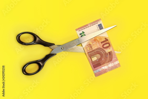 Tabletop view - large scissors cutting ten eur banknote over yellow board. Cut fees concept.