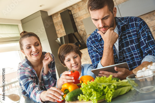 Family at home standing in kitchen together mother and son eating vegetbles while father working on digital tablet concerned photo